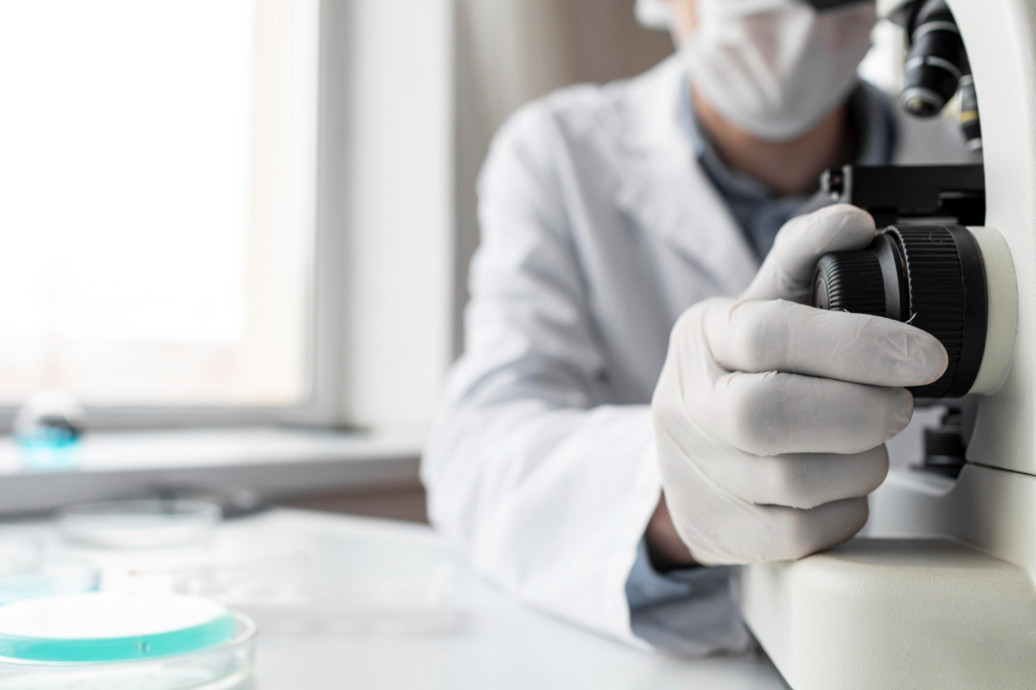A imagem mostra as mãos de uma pessoa sentada em um laboratório usando um microscópio. A pessoa veste jaleco branco, mascara branca N95 e usa luvas brancas de latex. A mesa é branca e tem um frasco transparente com líquido azul piscina dentro. Ao fundo, uma janela.
