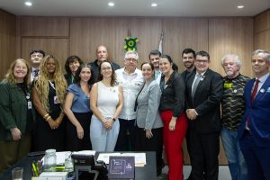 Pessoas em pé pousando para foto em um escritório com parede marrom de madeira. No centro da imagem, o ministro da saúde, Alexandre Padilha veste calça social preta e blusa social branca.