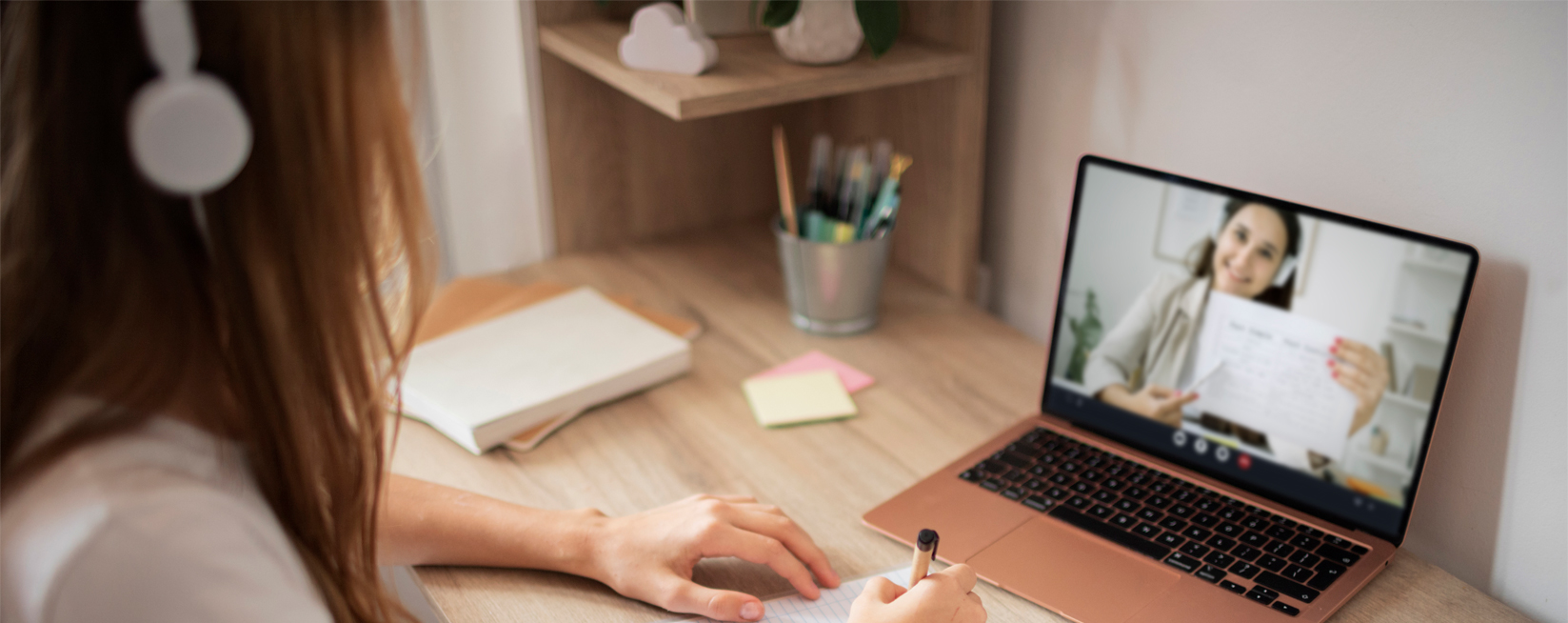 Mulher jovem de fios lisos e médios usando fone de ouvindo cinza sentada assistindo aula em um laptop preto.