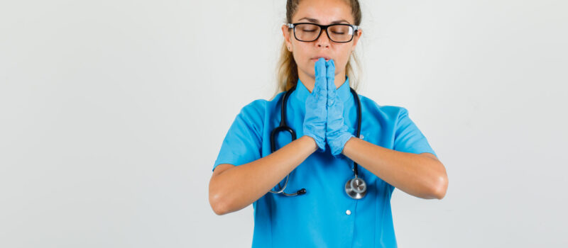 Médica-veterinária usando uniforme azul e óculos em pé com as mãos juntas em posição de oração e olhos fechados.