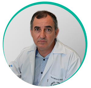 Homem branco por volta dos 40 anos sentado usando blusa social azul de botões no meio e jaleco branco. É o dr. Márcio Garcia Ribeiro.